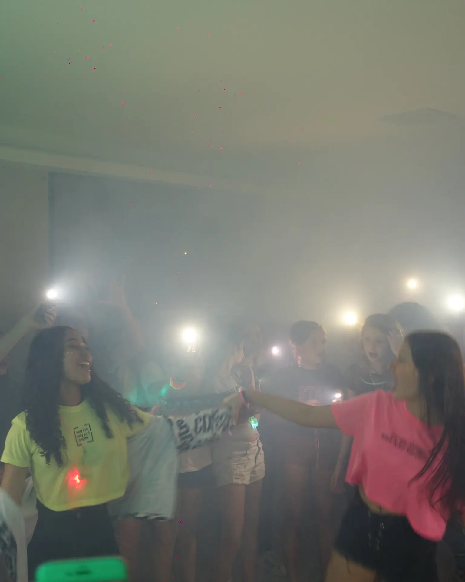 Adolescentes cantando e dançando na pista com fumaça e luzes de celular.
