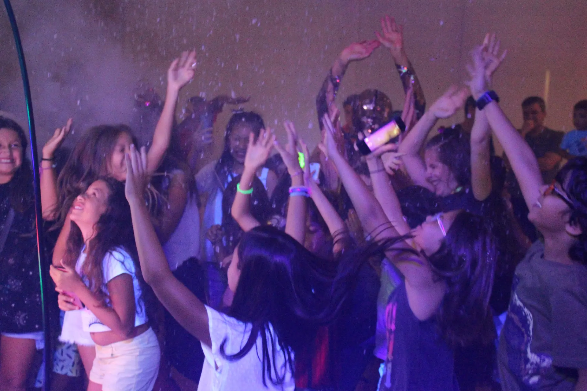 Adolescentes com os braços erguidos na pista de Bar e Bat Mitzvah sob luzes neon e chuva de papel.