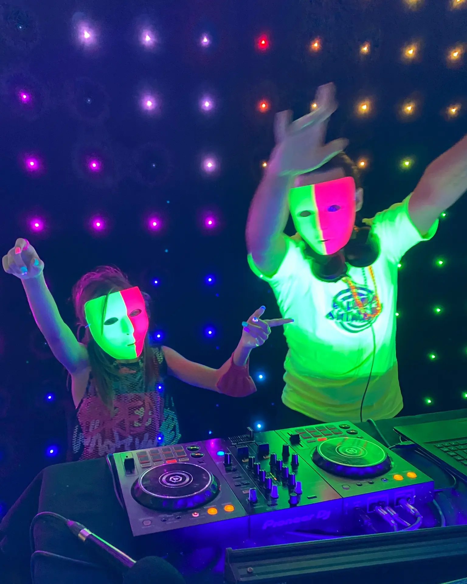 Crianças com máscaras neon dançando ao lado da controladora do DJ em festa infantil.