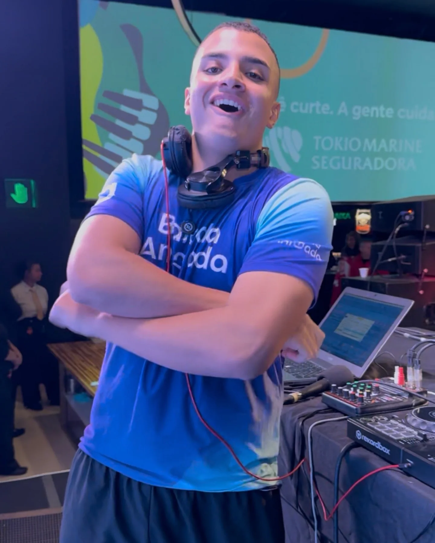 DJ da Balada Animada em evento corporativo com telão da marca ao fundo.