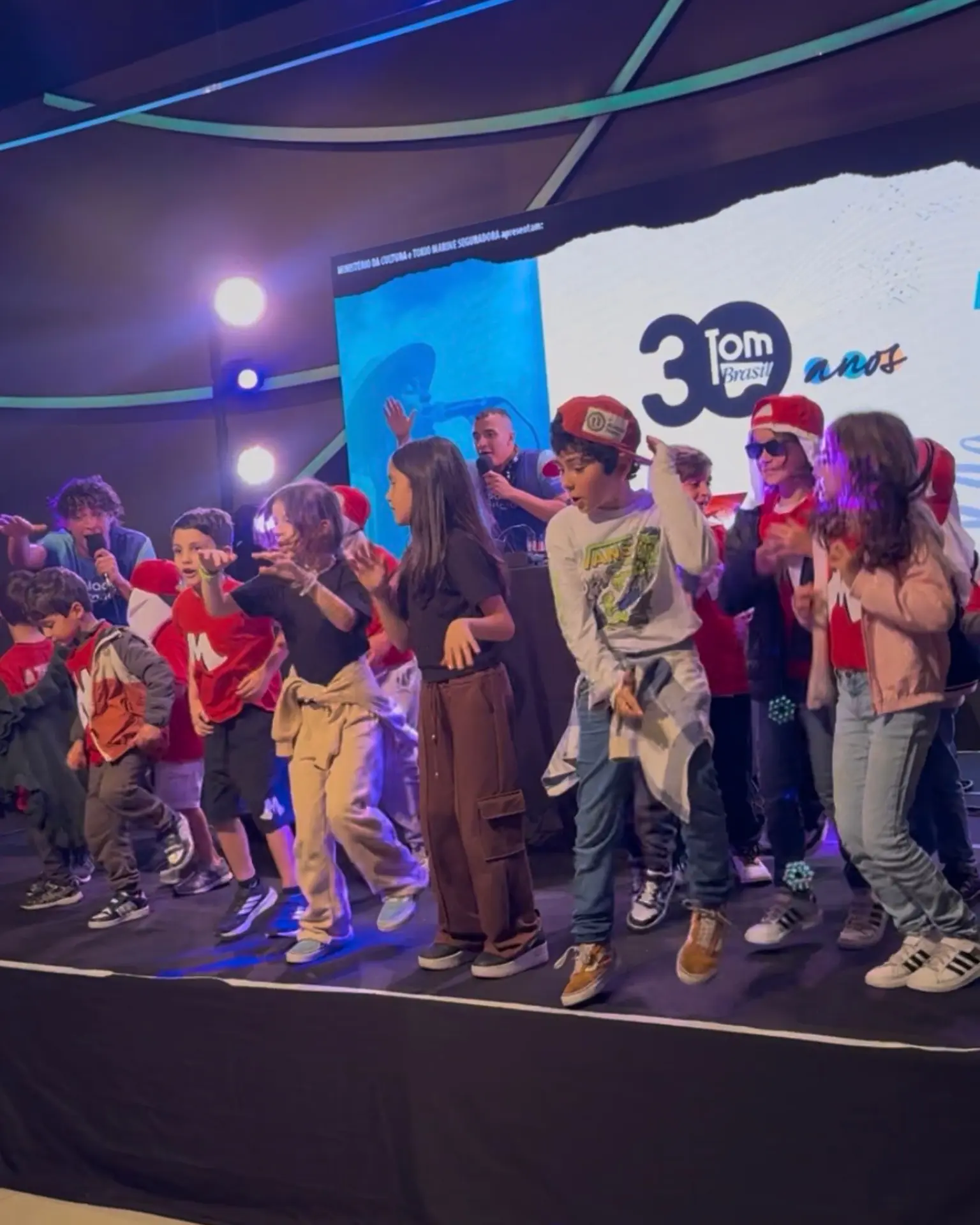 Crianças dançando no palco de um espaço de shows com o DJ ao microfone.